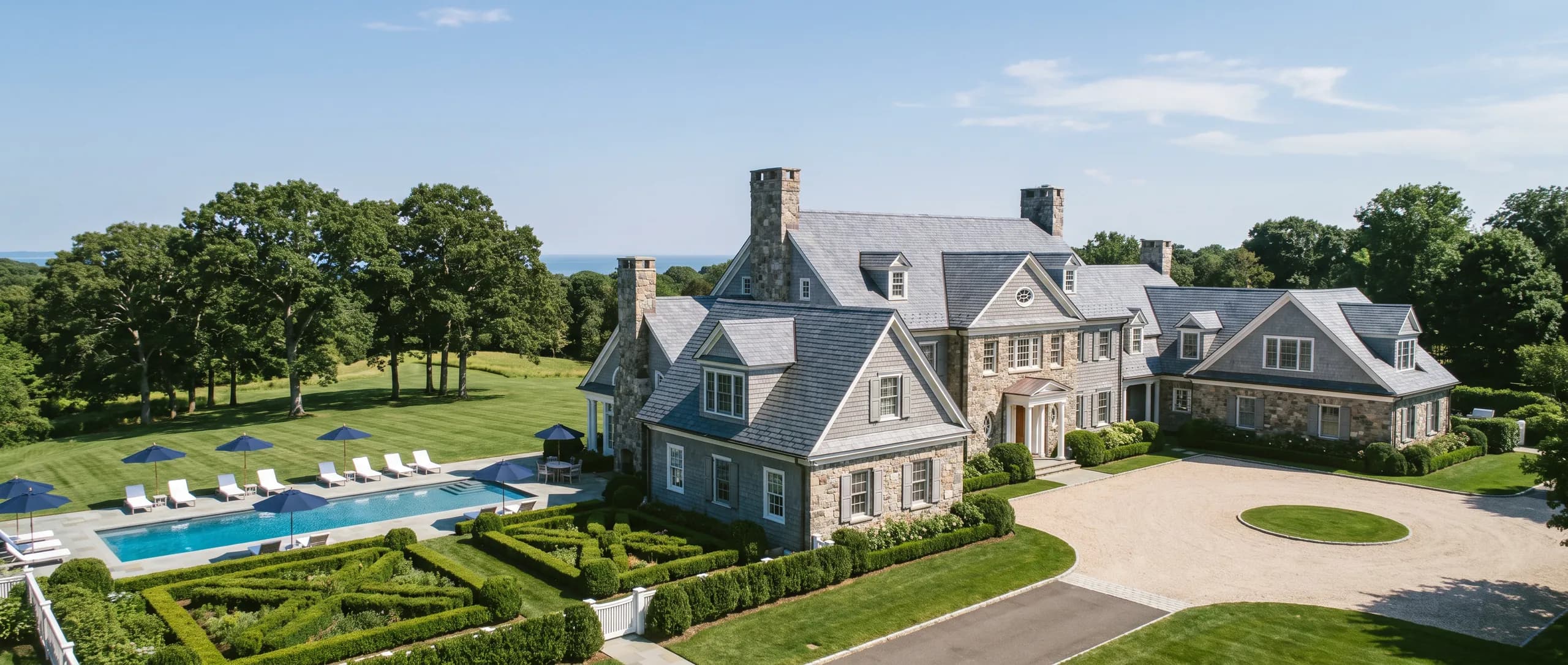 An elevated summer view of a shingle and stone estate with a long pool, white chaises, boxwood parterres and lawn rolling to old oaks.